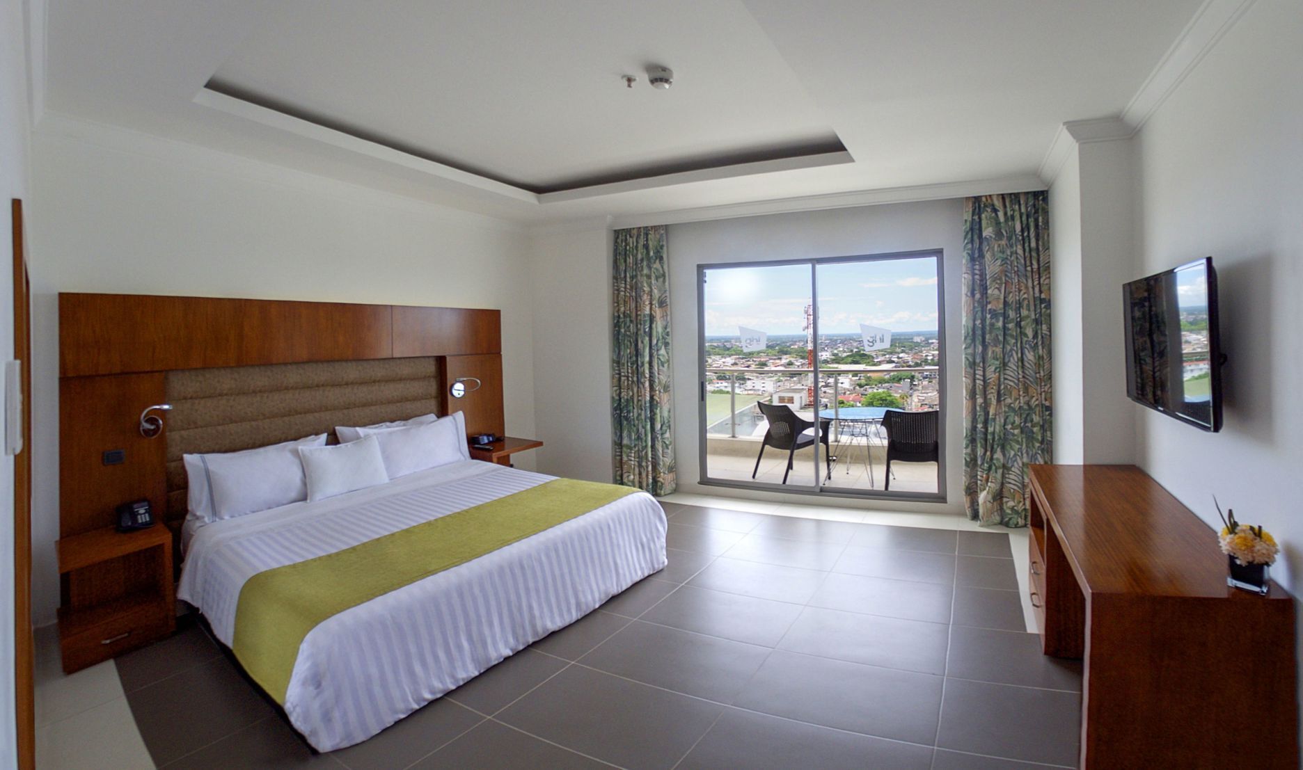 A hotel room with a bed, a TV, a balcony with a table and chairs, and a view of the city.