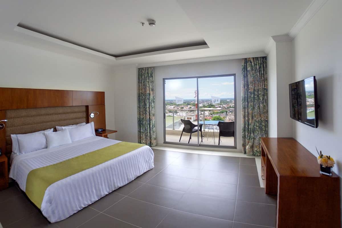 Une chambre d’hôtel avec un lit, une télévision, un balcon avec une table et des chaises, et une vue sur la ville.