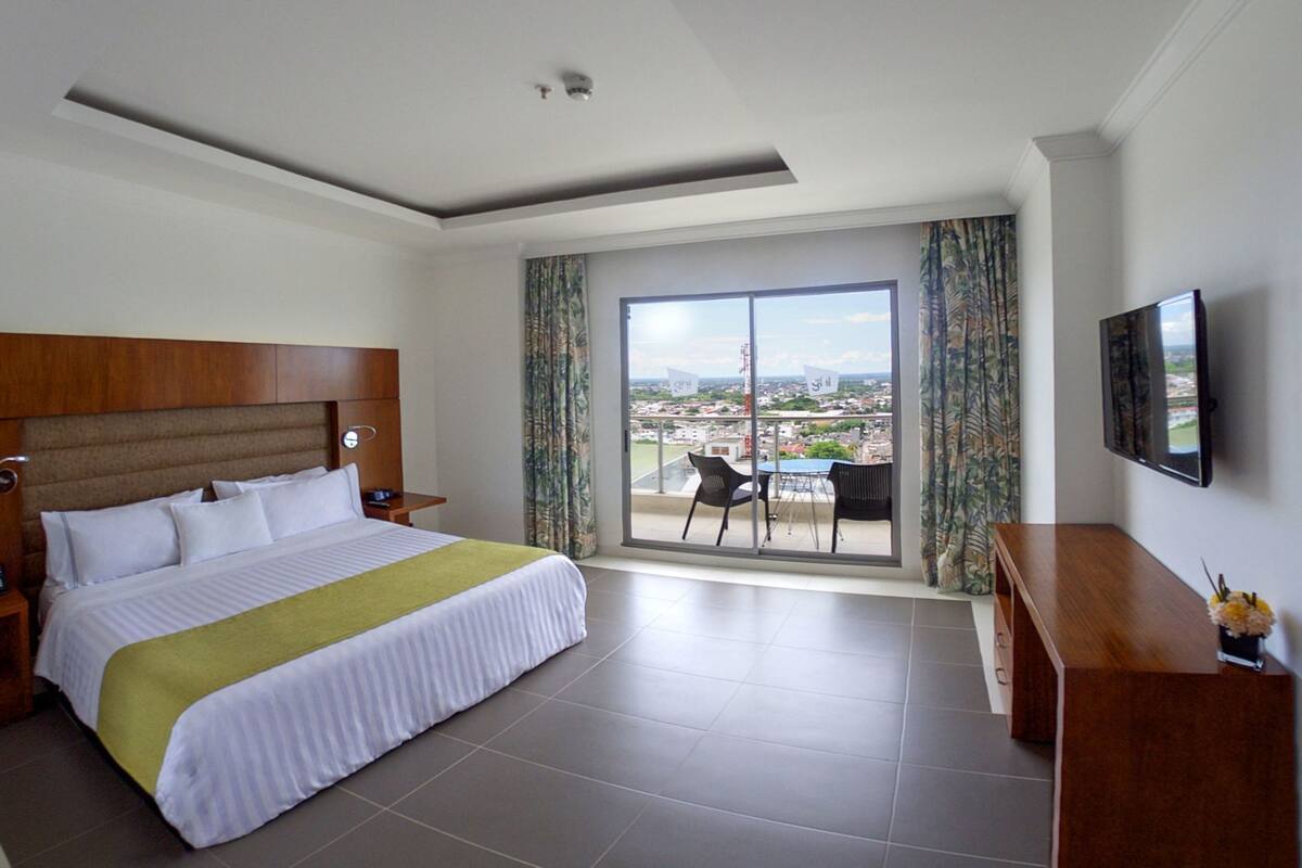 Habitación de hotel con cama, televisor, balcón con mesa y sillas, y vista a la ciudad.