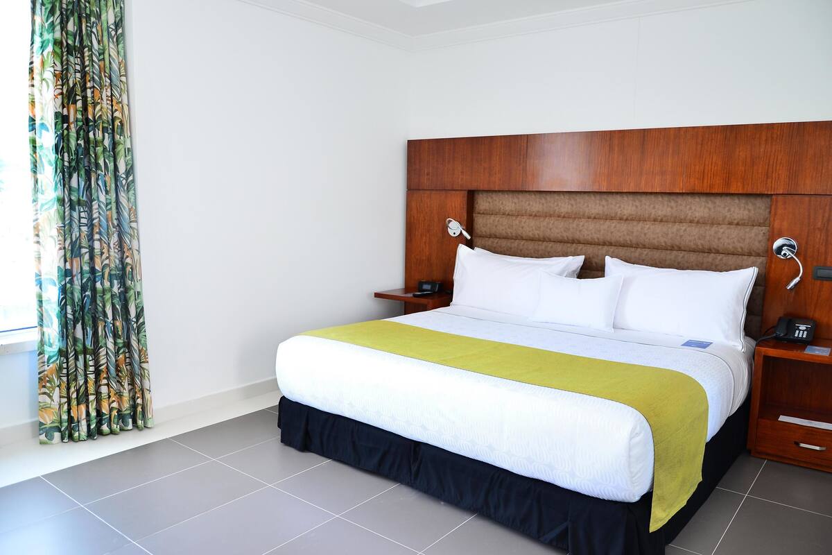 Une chambre d’hôtel avec un grand lit, une tête de lit en bois, deux tables de chevet et un rideau orné d’un motif de feuilles.