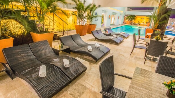 Un espace aménagé au bord de la piscine, avec des chaises longues et des tables, entouré de plantes en pot et donnant sur une grande piscine intérieure.