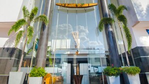 Un hôtel moderne à la façade blanche, doté de plusieurs étages et d’une entrée imposante avec des portes en verre et des palmiers.