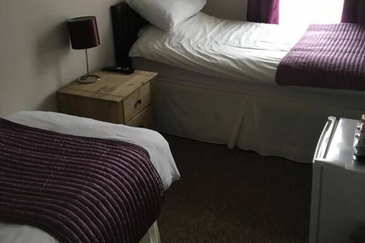A small, dimly lit room with two single beds, a nightstand, a desk, and a window with curtains.