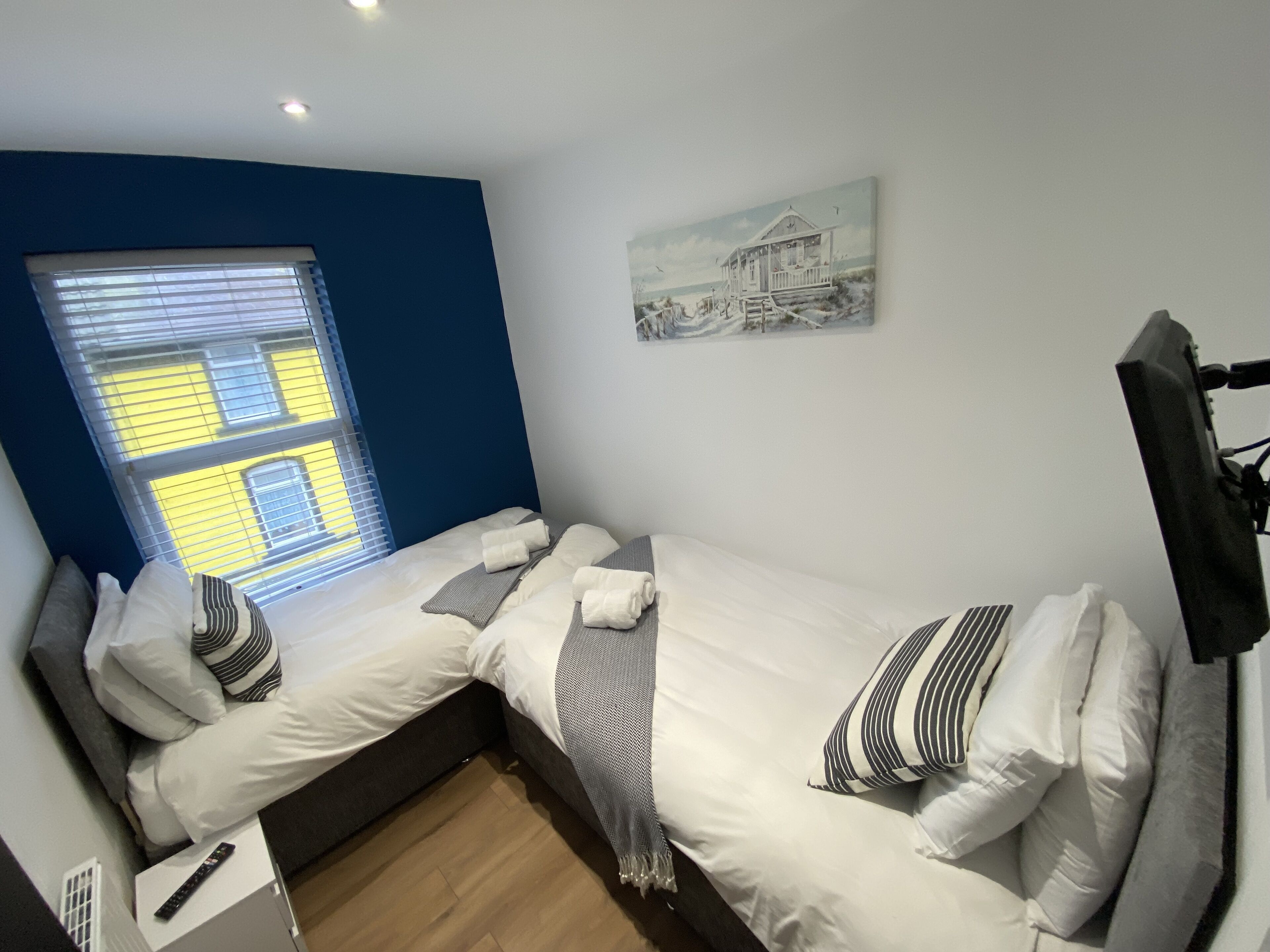 Ein kleiner, gemütlicher Raum mit zwei Betten, einem Fenster mit Jalousien, einem Bild an der Wand und einem an der Wand befestigten Fernseher.
