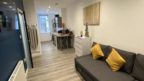 A modern living room with a grey sofa and yellow cushions, a kitchen area in the background, and a large window providing natural light. - Blackpool Abode - 20 Bairstow Street (Blackpool)