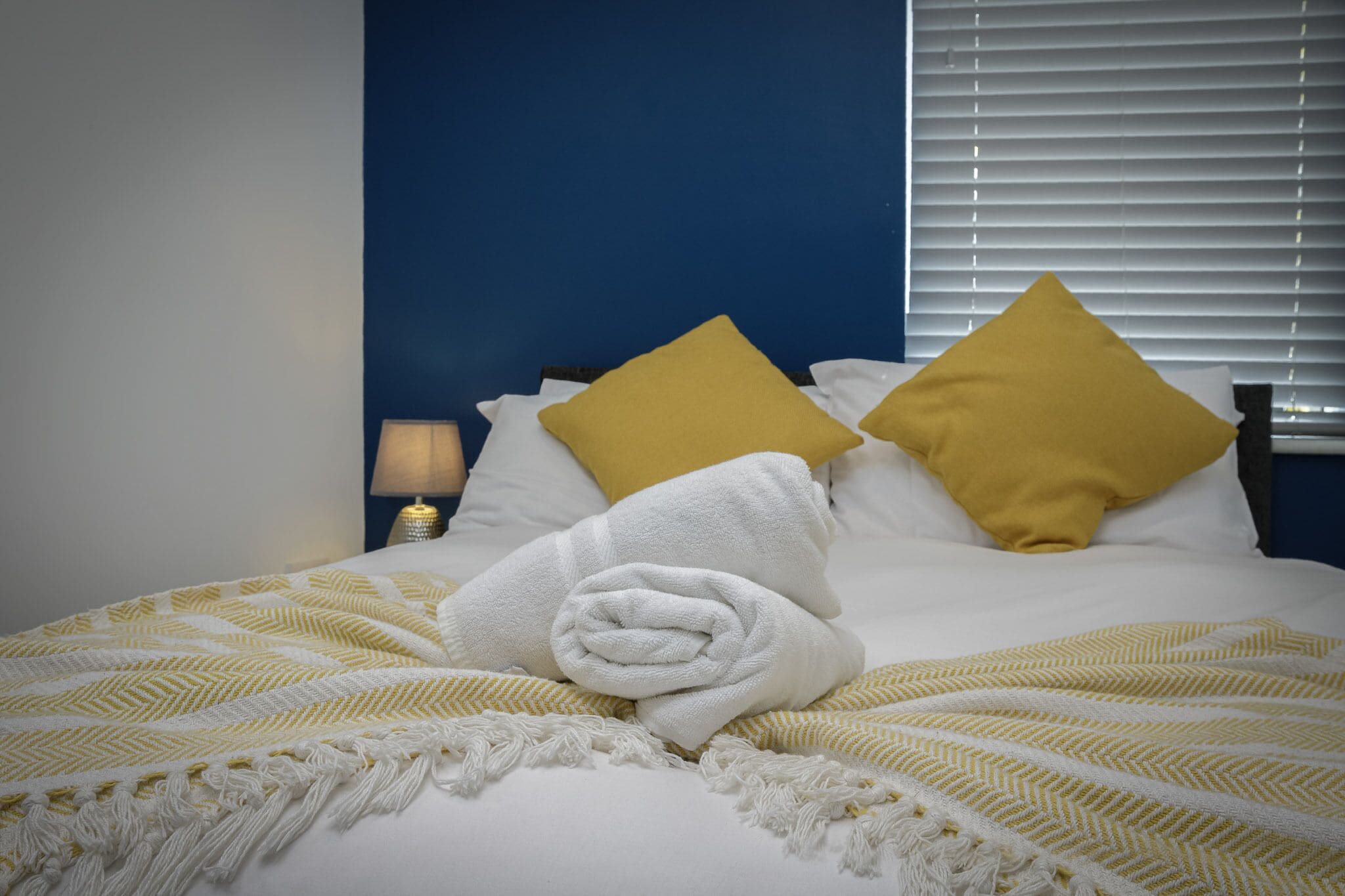 A neatly made bed with a rolled white towel, two mustard-colored pillows, and a striped blanket.