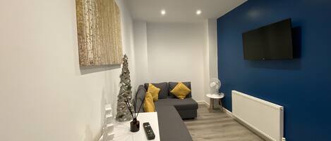 A modern living room with a sectional sofa, a small Christmas tree, and a flat-screen TV mounted on the wall.