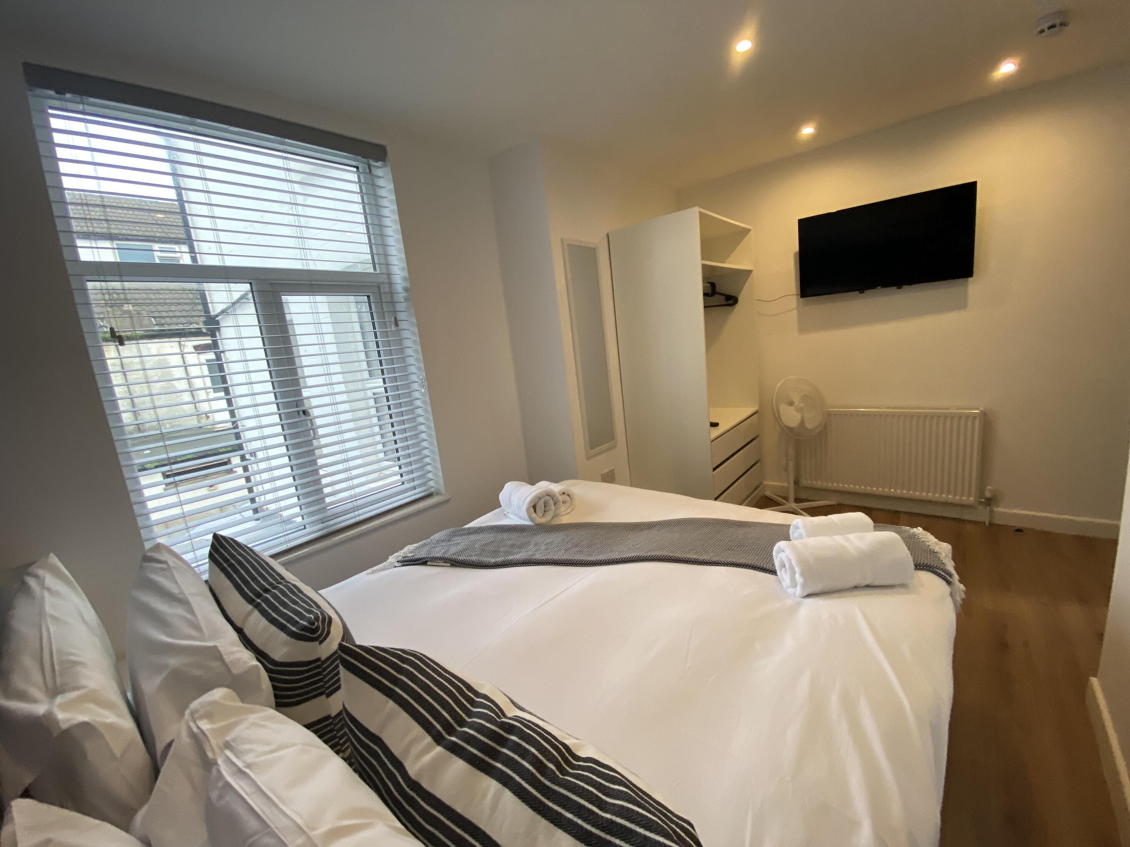 A hotel room with a bed, a window with blinds, a wardrobe, a radiator, and a flat-screen TV mounted on the wall.