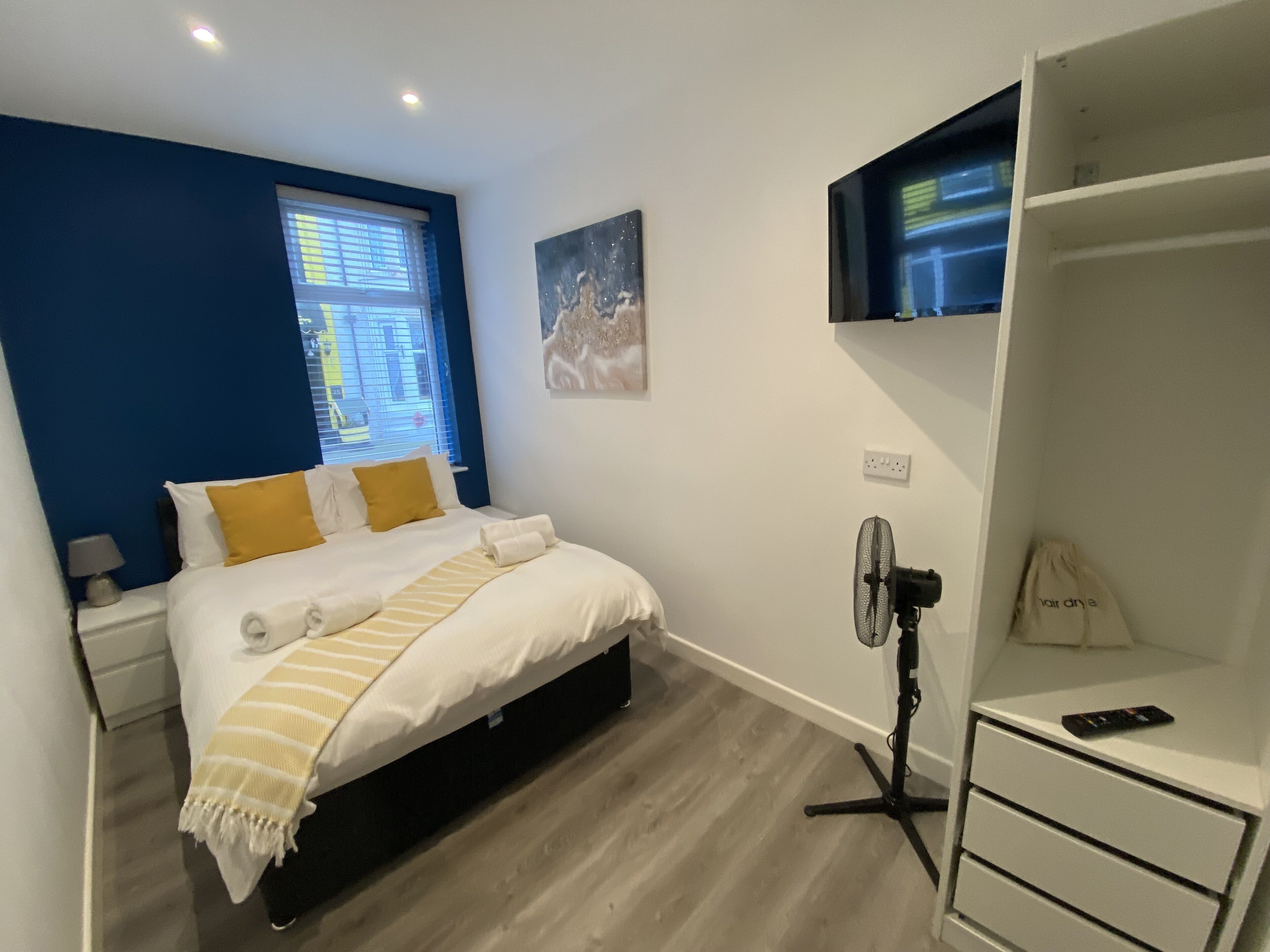 A modern bedroom with a bed, a standing fan, a TV on a stand, and a window with blinds.