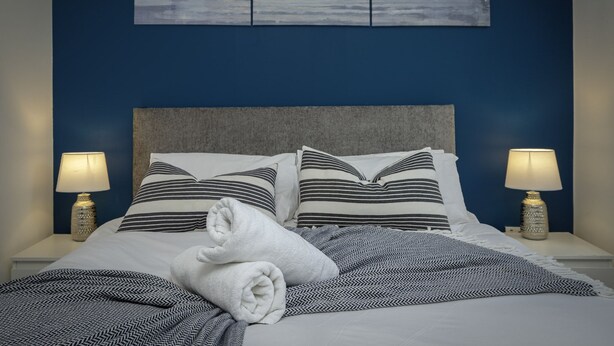 A neatly made bed with striped pillows and a rolled towel, two bedside lamps, and a painting of a coastal scene on the wall. - Blackpool Abode - 20 Bairstow Street (Blackpool)