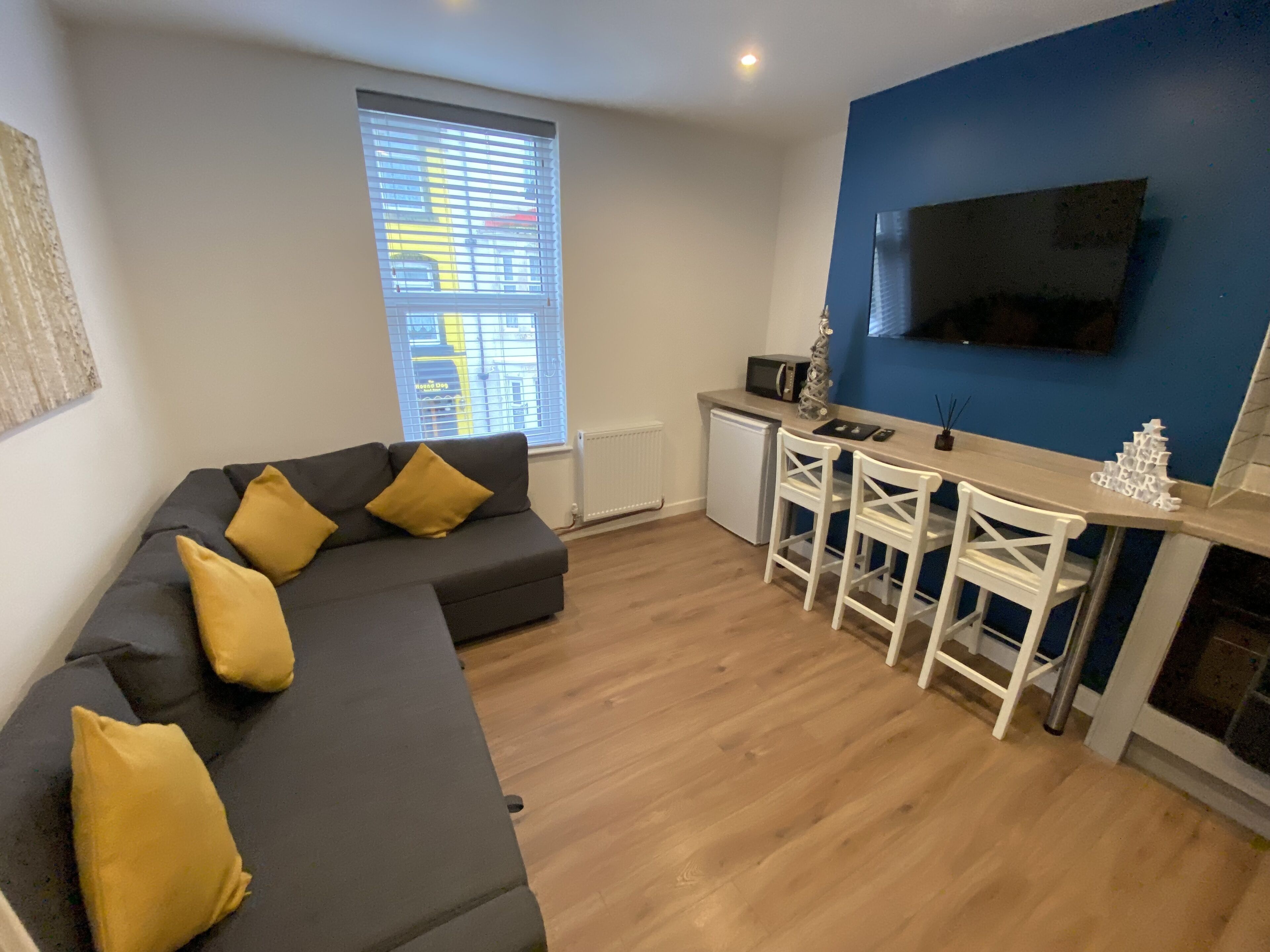 A modern living room with a sectional sofa, a small kitchen area, and a TV mounted on the wall.