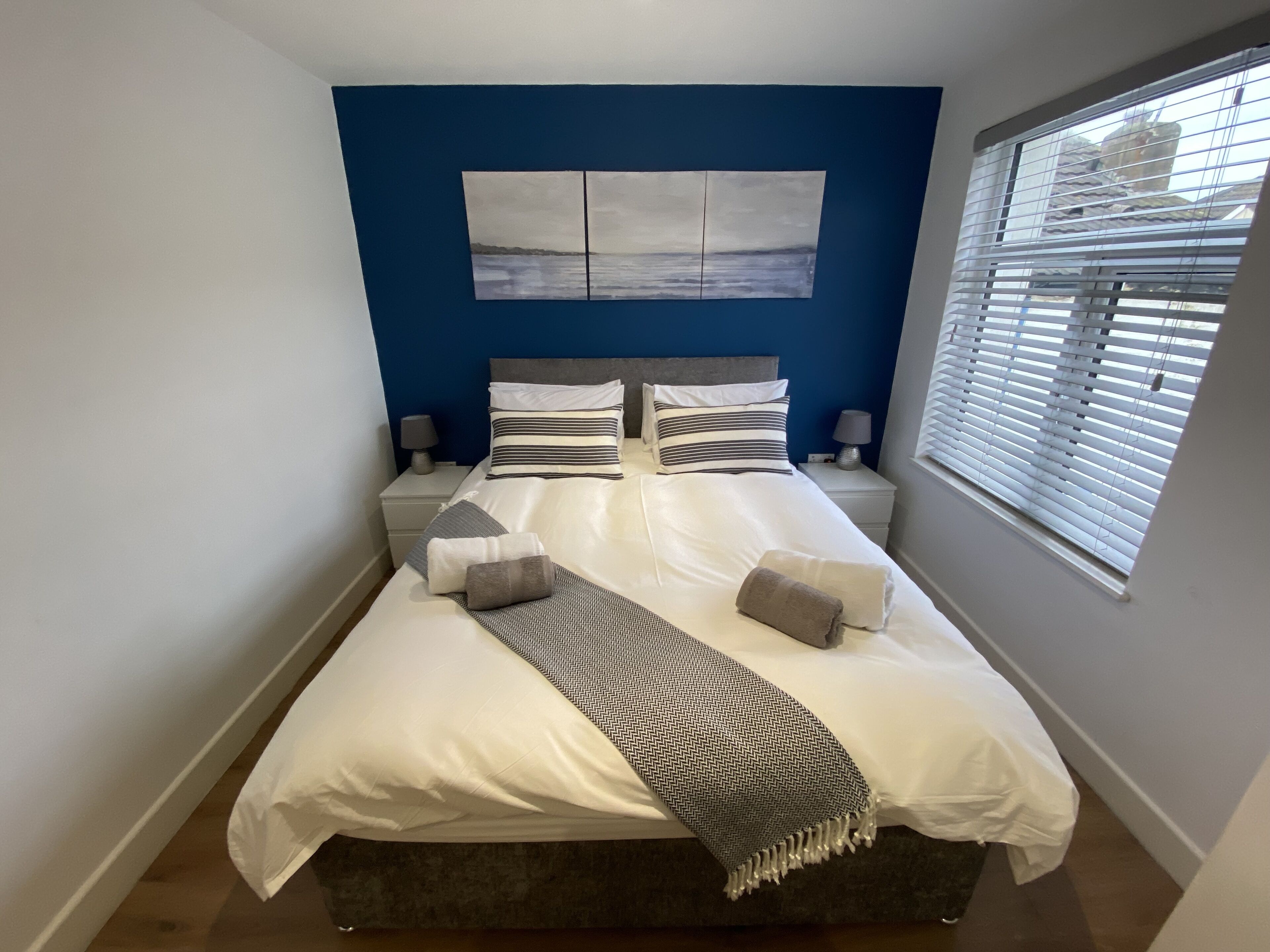A neatly made bed with a white comforter and multiple pillows, a blue accent wall, a window with blinds, and a small bedside table with a lamp.