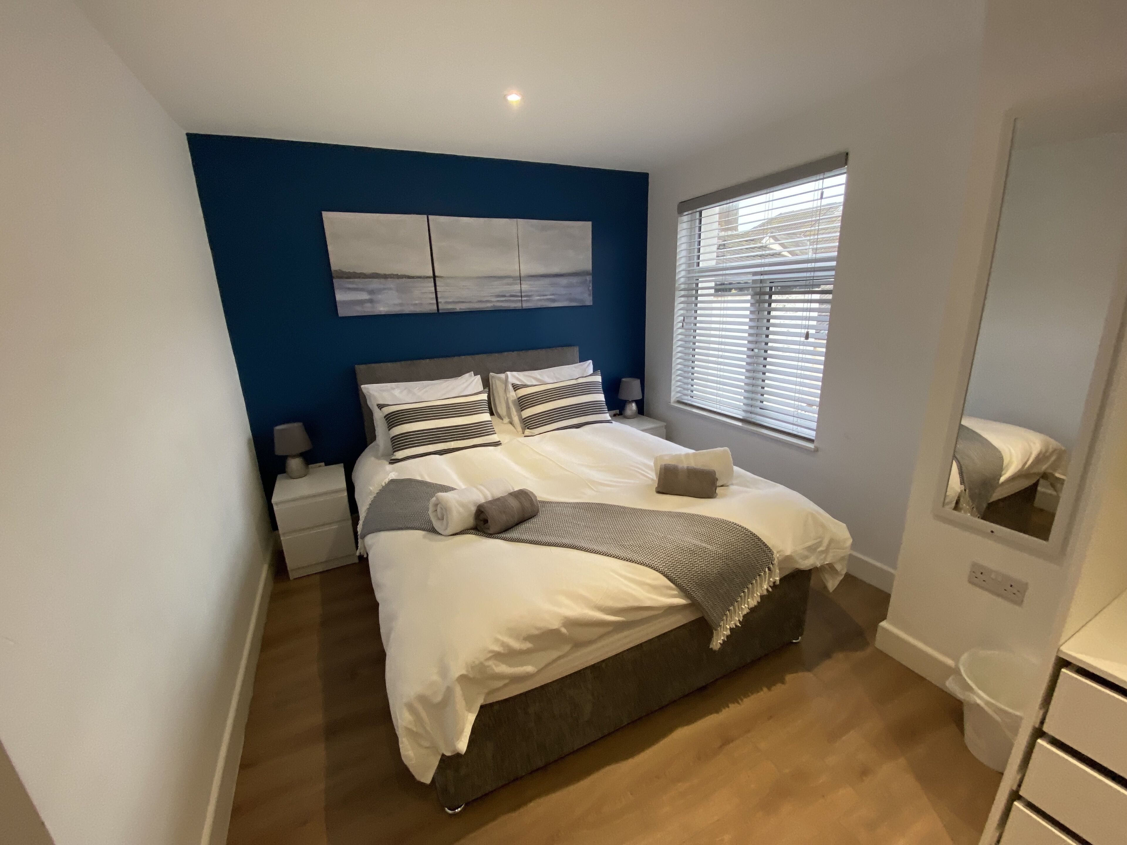 Une chambre moderne avec un grand lit, une table de chevet, une commode, une fenêtre avec des stores et une œuvre d’art accrochée au mur.