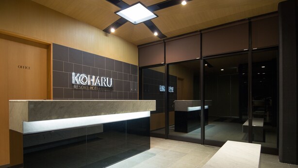 A reception area with a modern design, featuring a reception desk, a sign for Koharu Resort Hotel, and a glass door leading to the interior. - Koharu Resort Hotel & Suites (Hakuba)