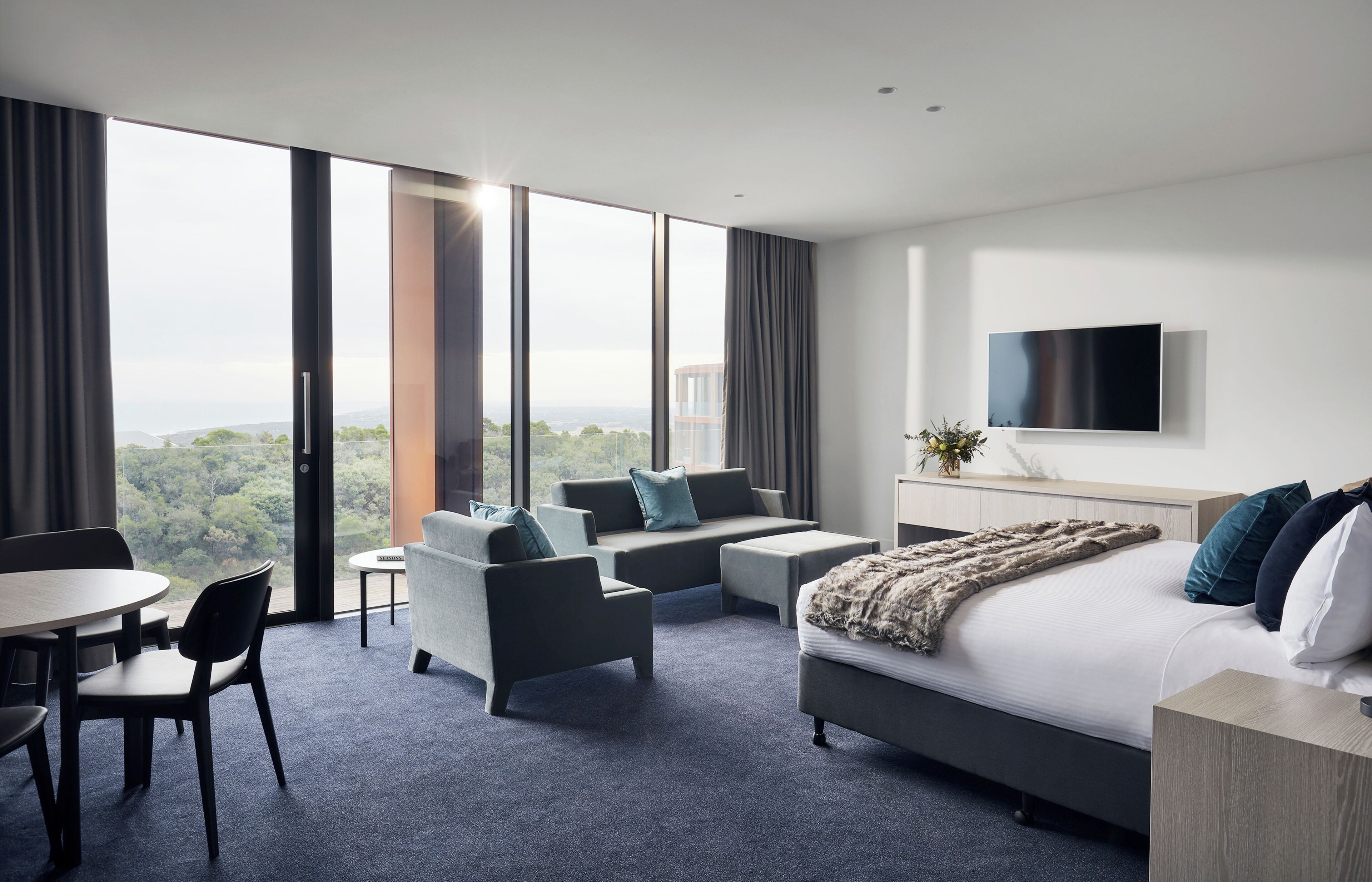 A modern hotel room with a large bed, a sofa, a dining table, and a TV.