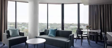 A modern hotel room with a large window, a sofa, a round table, and a small dining area.
