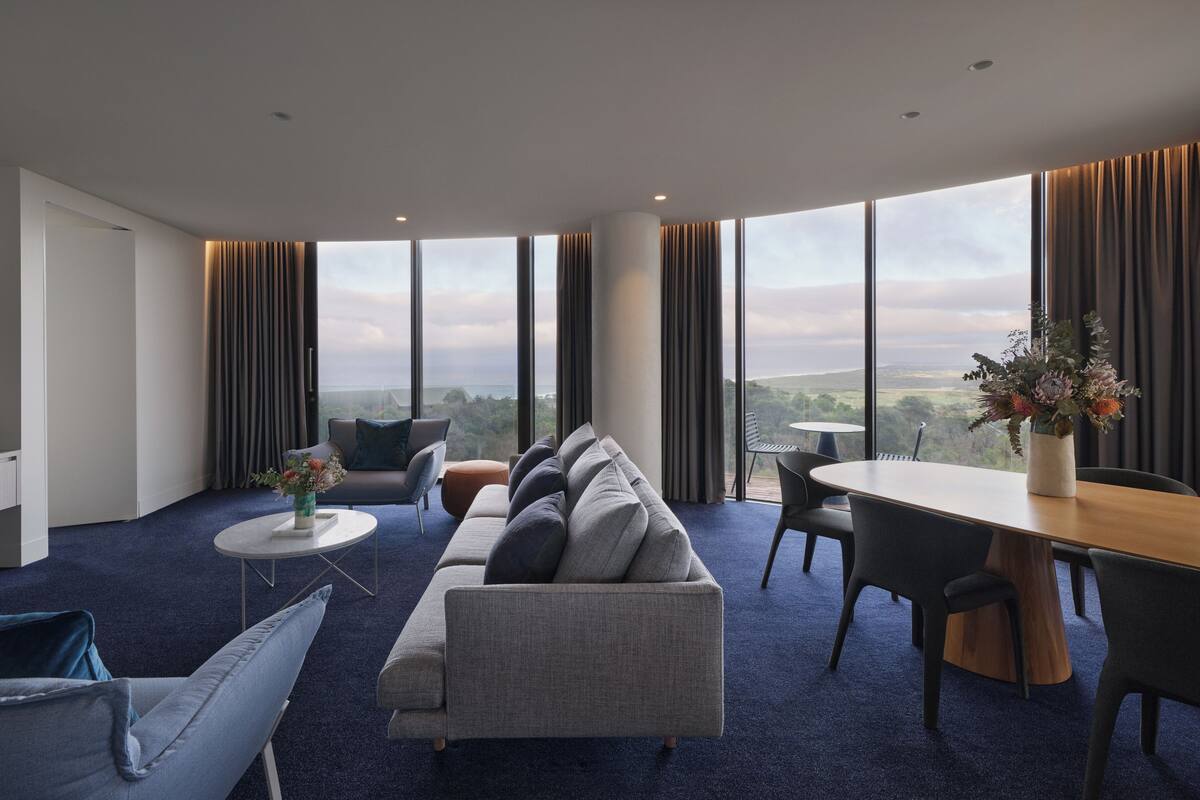 Habitación de hotel moderna con un amplio ventanal que ofrece vistas a la ciudad, un conjunto de sofás, una mesa de comedor y una pequeña mesa de centro.