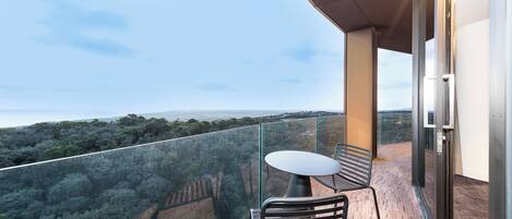 A balcony with a glass railing, a table, and two chairs, overlooking a forested landscape.
