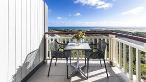 Ein Balkon mit weißer Brüstung, zwei schwarze Netzsessel, ein weißer Tisch und eine Vase mit Blumen.