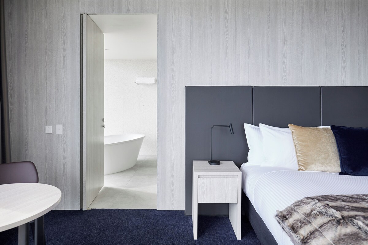 A modern hotel room with a bed, bedside table, and a bathroom visible through an open door.