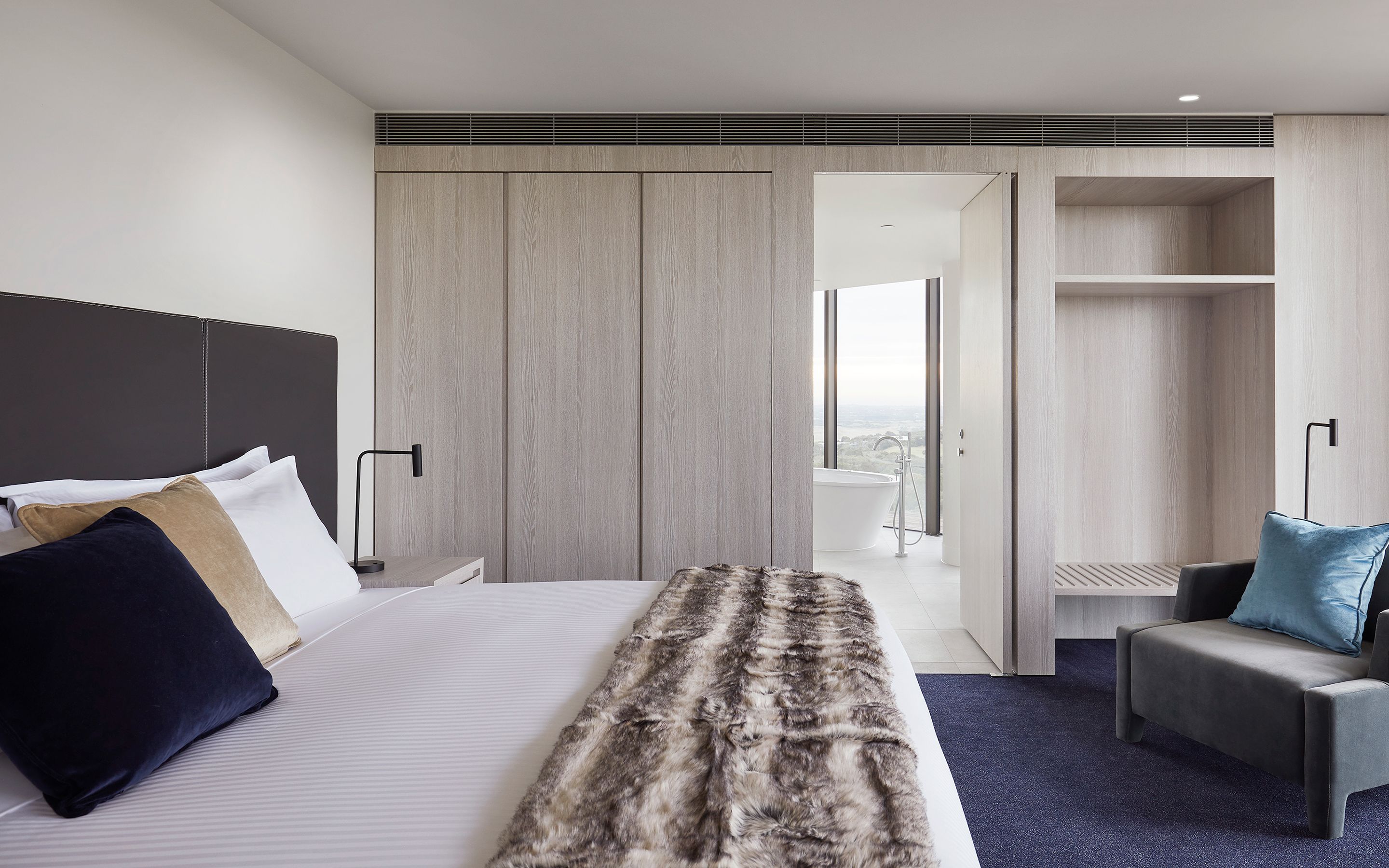 Ein modernes Hotelzimmer mit einem großen Bett, einer Bank, einem Schreibtisch und Blick ins Freie.