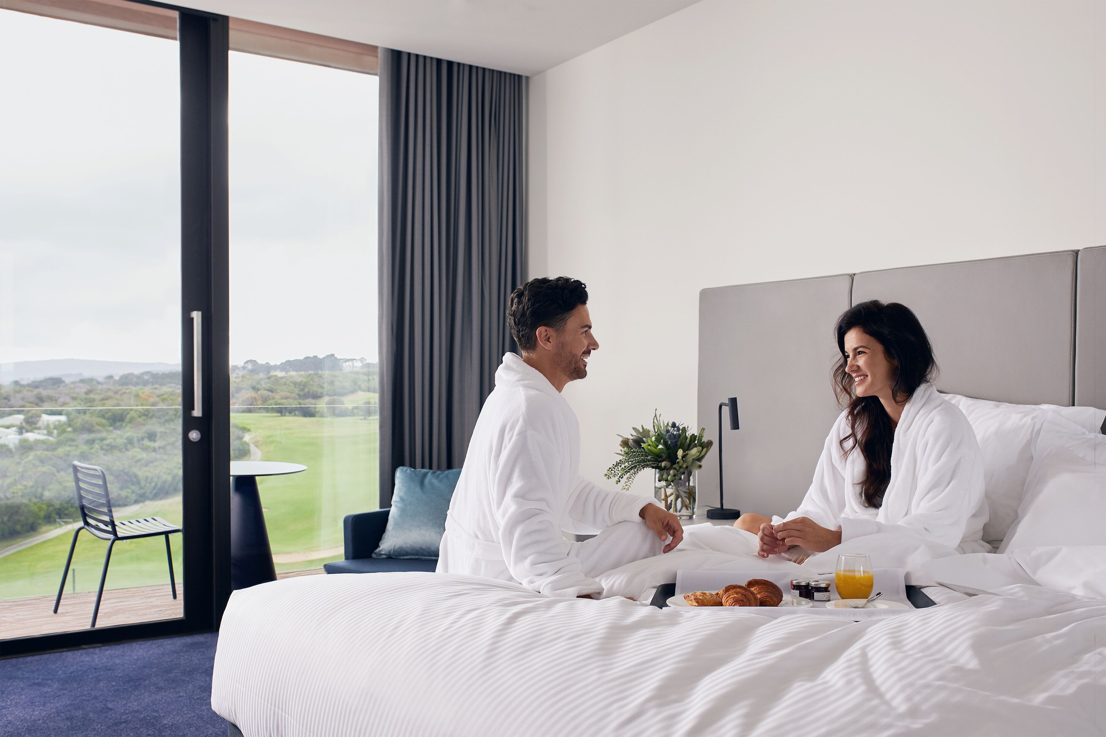 A hotel room with a large bed, a dining table, and a view of a green landscape.