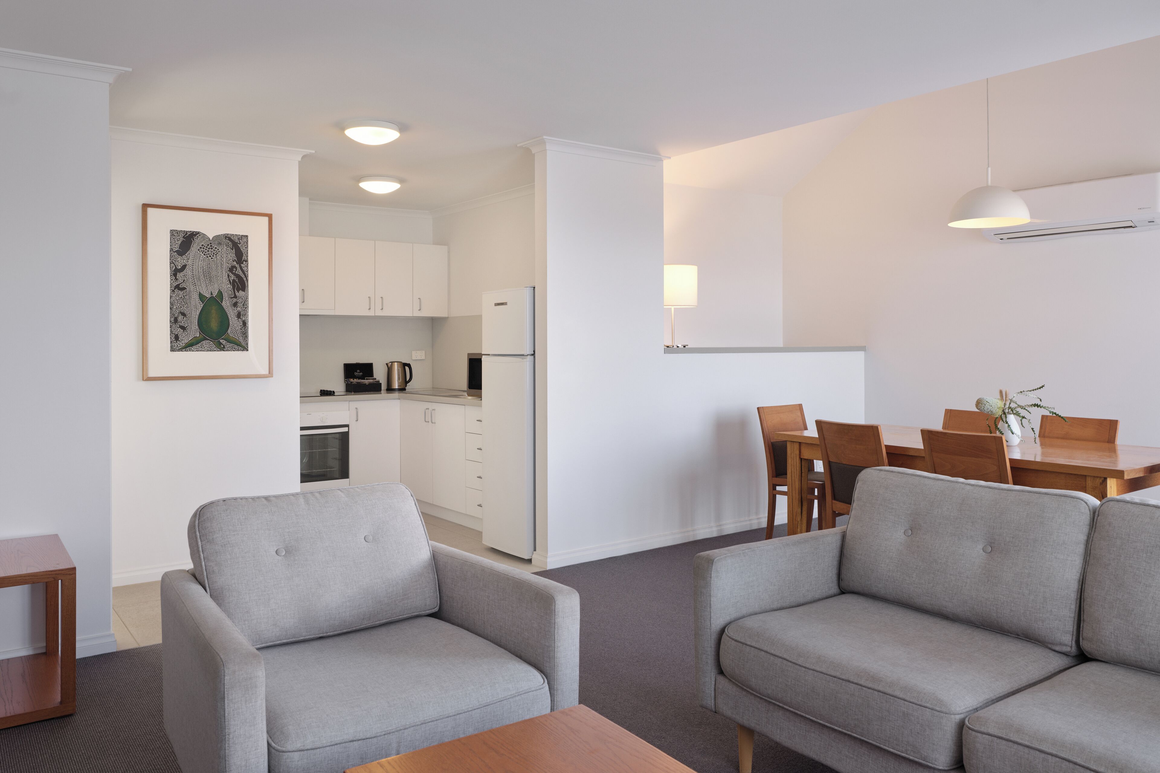 A modern living room with grey sofas, a wooden coffee table, and a dining area with a table and chairs. A kitchen is visible in the background.