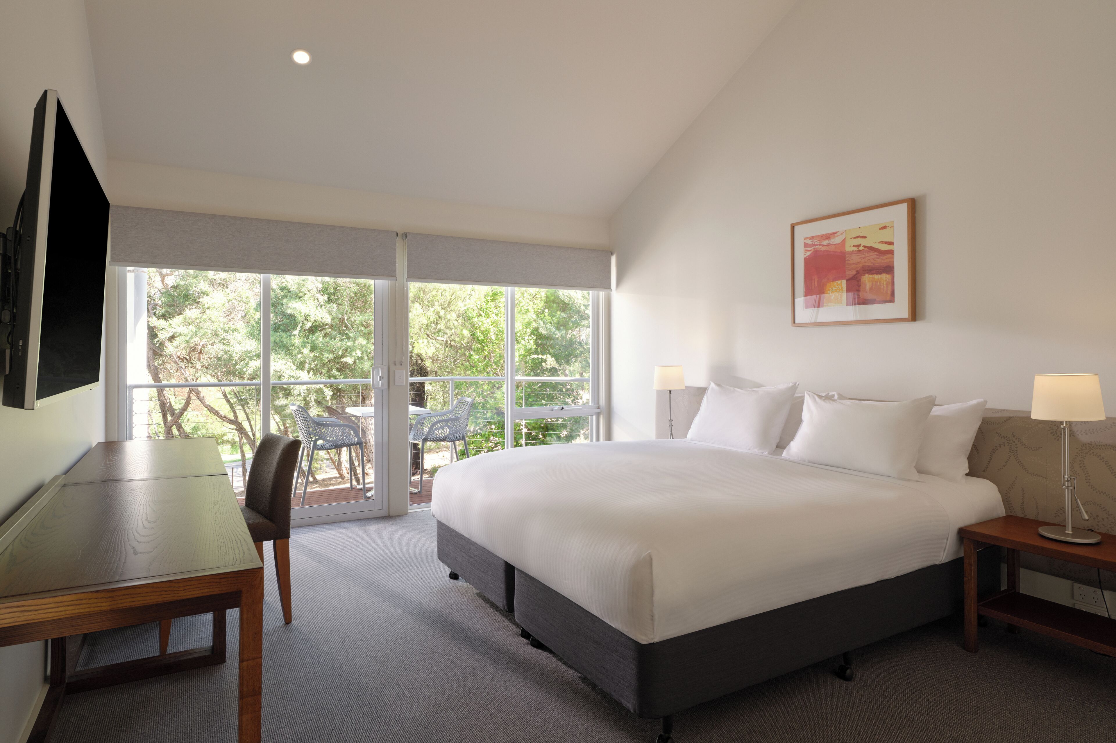 A hotel room with a large bed, a desk, and a chair. There is a window with a view of trees and a balcony.
