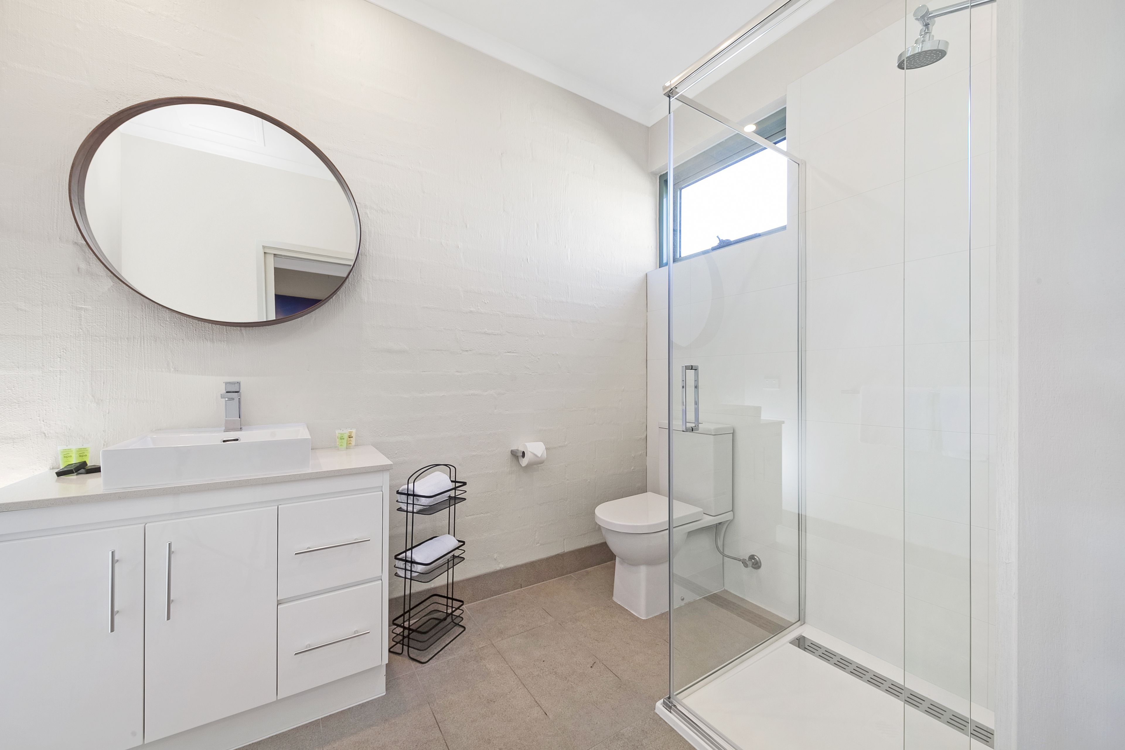 Un baño moderno con cabina de ducha de vidrio, un espejo redondo y un lavabo blanco con toallero.