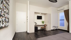 Una habitación de hotel moderna con suelo de madera oscura, una cama, un escritorio con silla, un televisor montado en la pared y una ventana con cortinas.