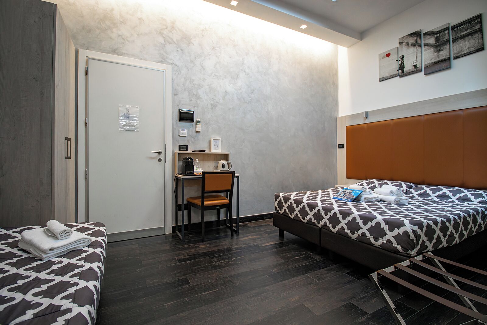 A hotel room with two beds, a desk, and a chair. There is a door, a small shelf with items, and a wall with artwork.