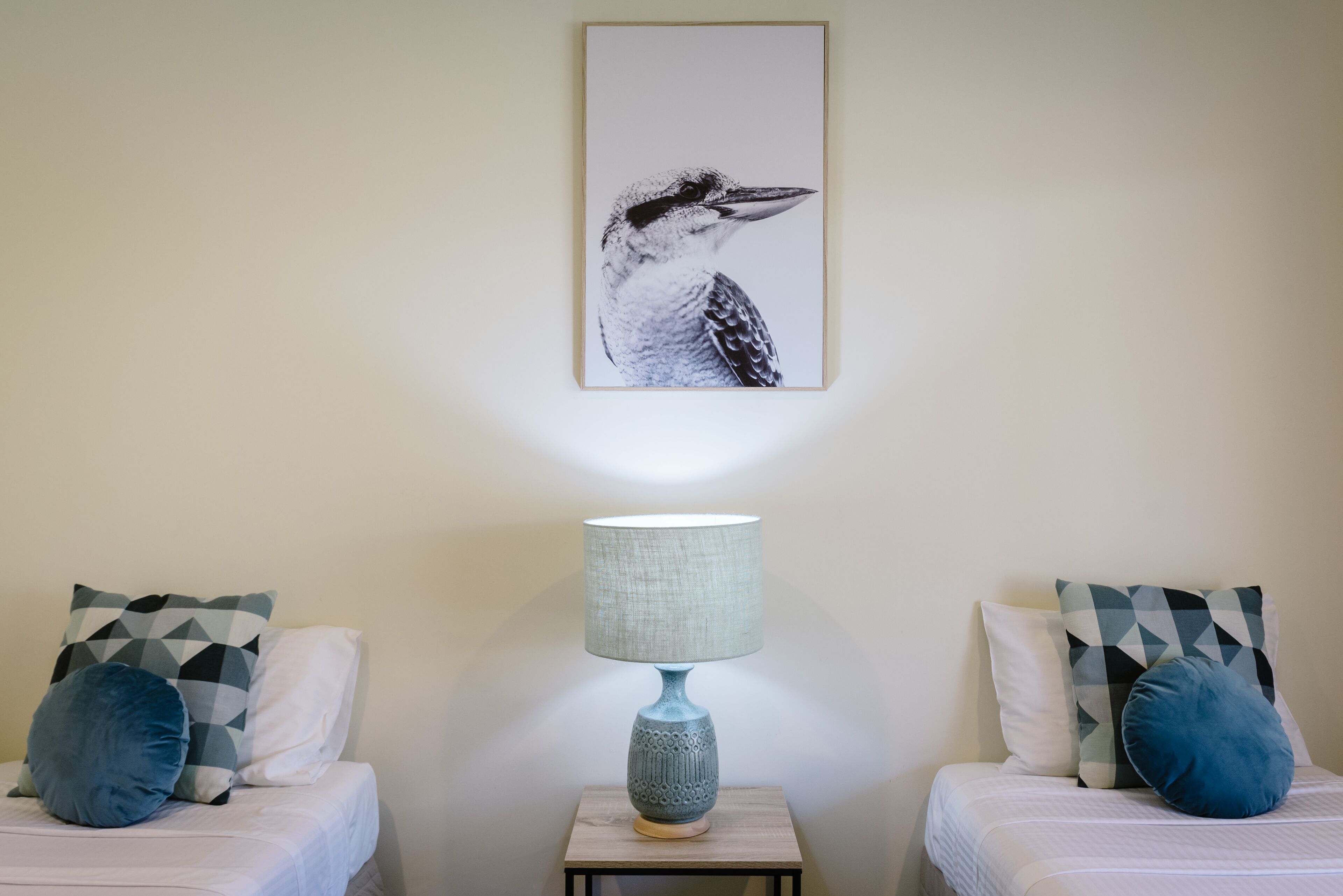 A bedroom with two beds, a nightstand with a lamp, and a framed bird artwork on the wall.