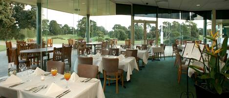 A restaurant with tables set for dining, large windows offering a view of a golf course, and a green carpeted floor.