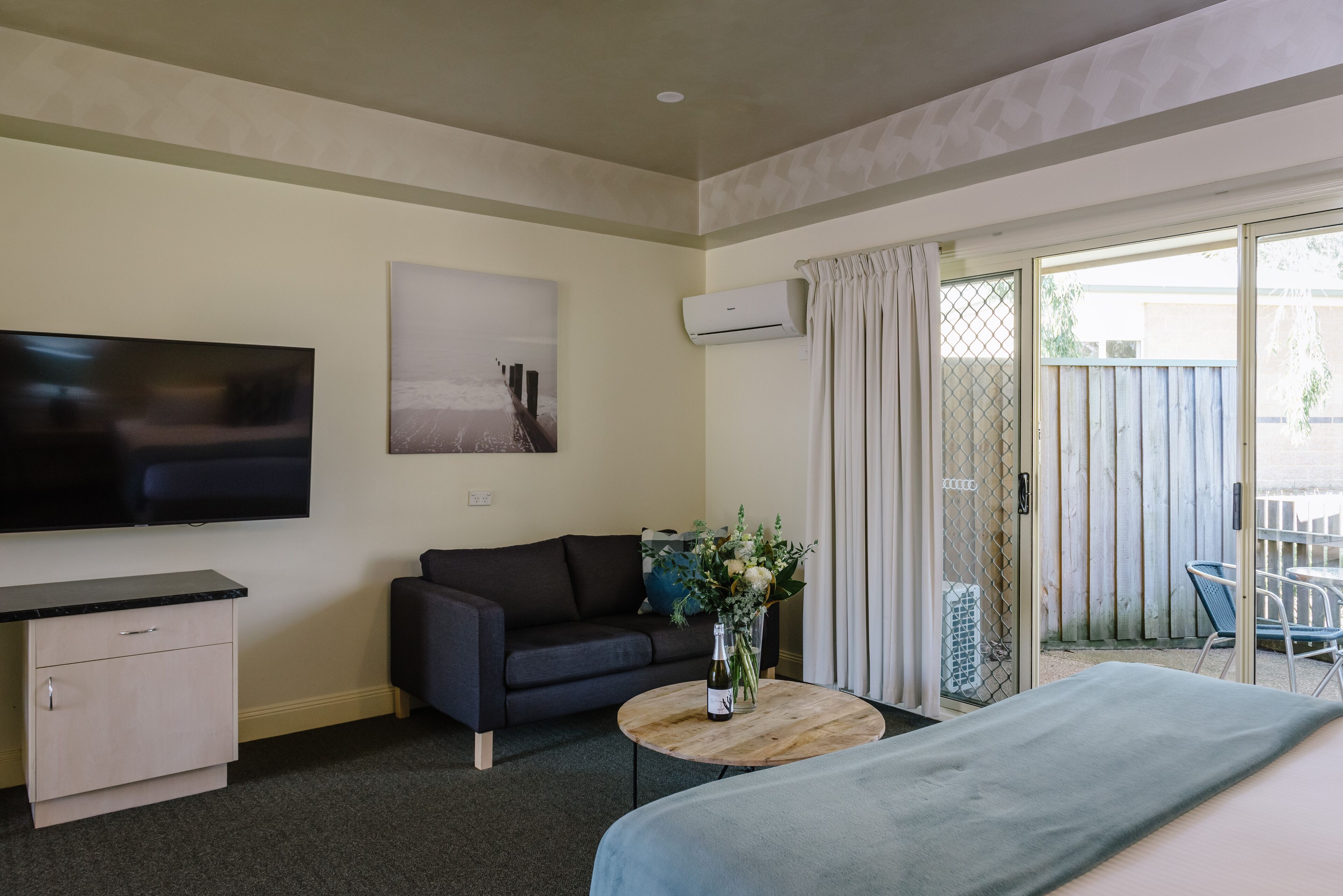 Una habitación con cama, sofá, televisión, una mesita con un florero y un ventanal corredero que da a un espacio exterior.