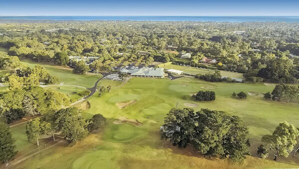 An aerial view of a golf course with a clubhouse, surrounded by dense greenery and residential areas.