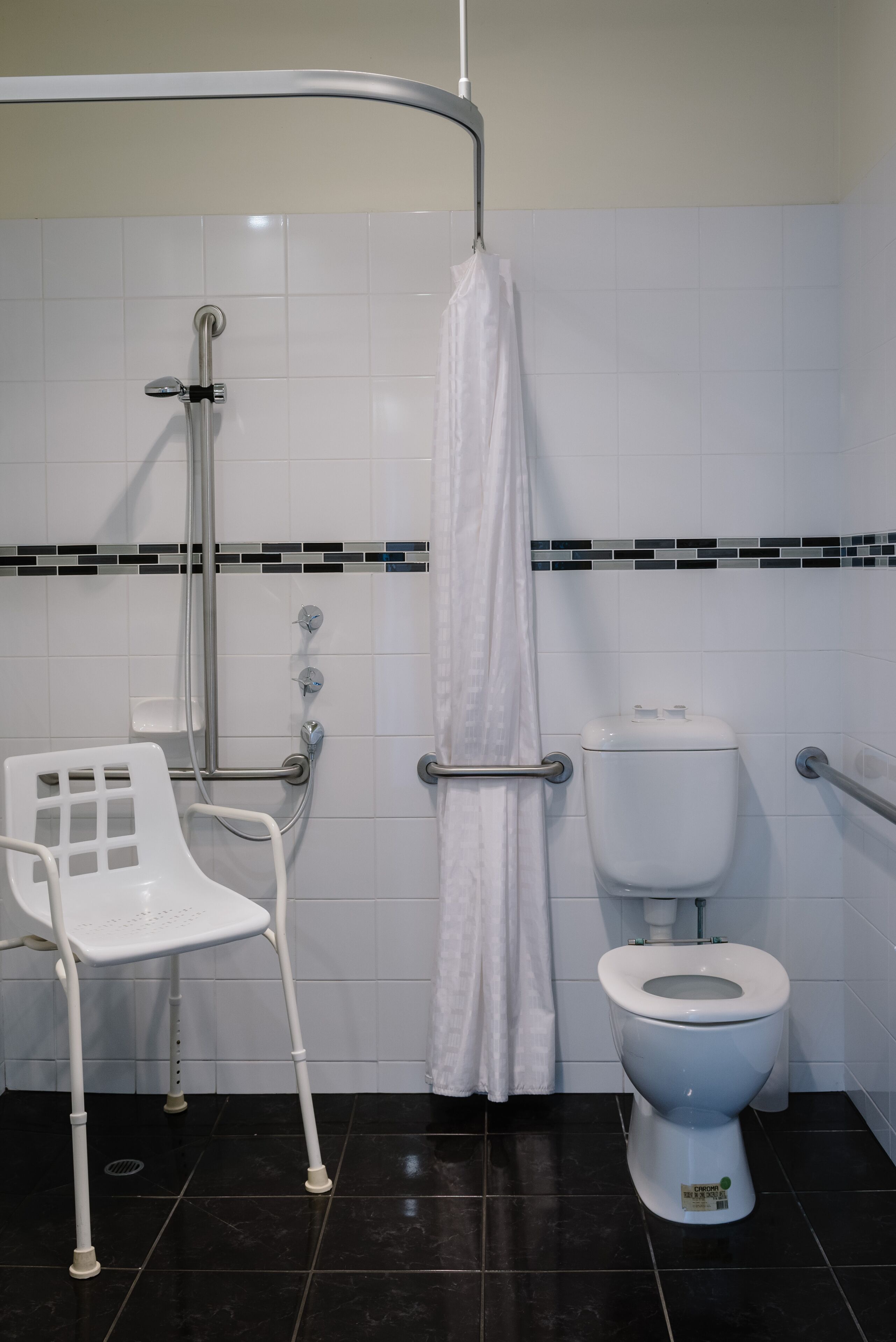 Baño con inodoro blanco, asiento de ducha y barra de apoyo.