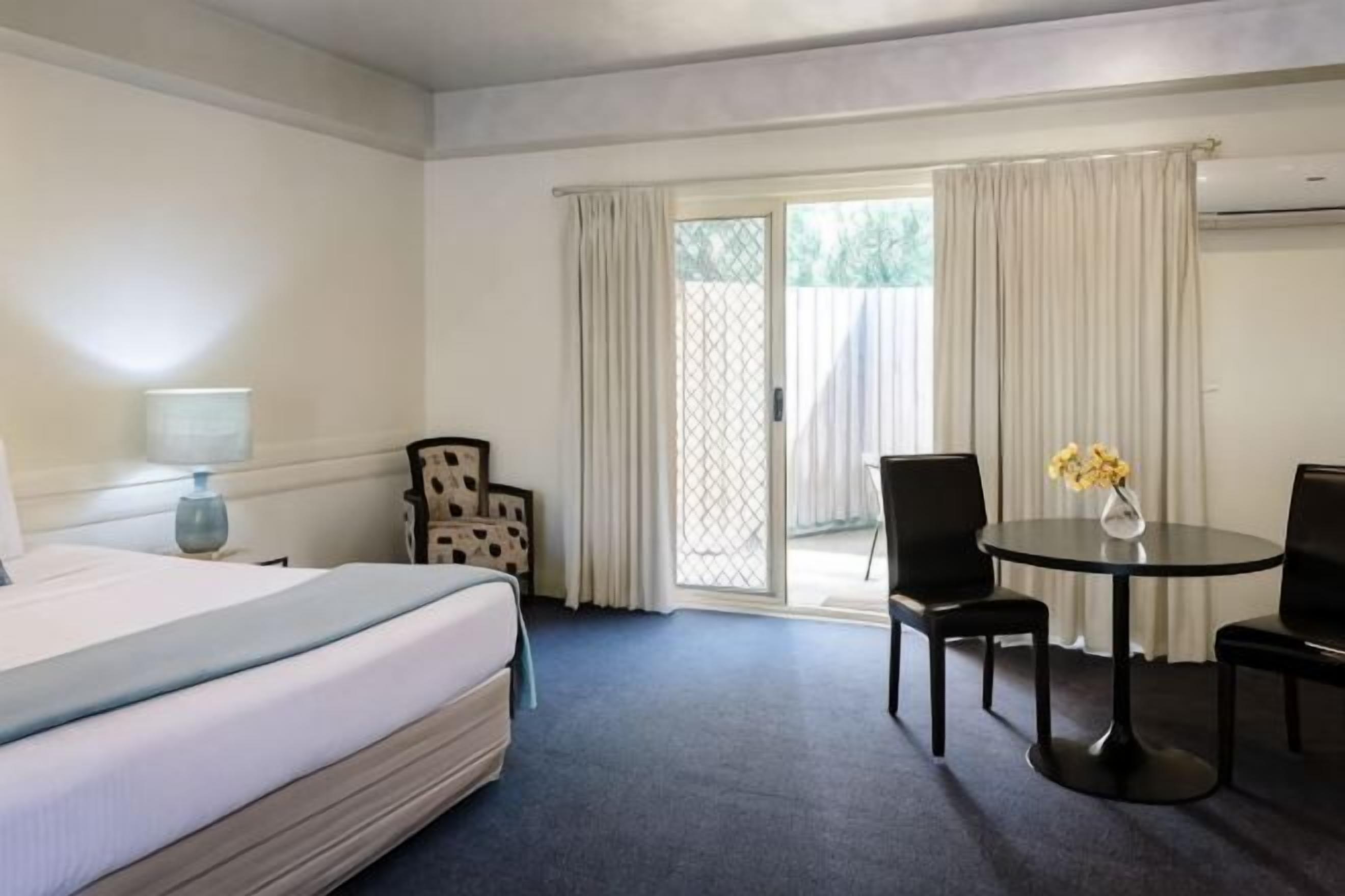 Habitación de hotel con cama, escritorio, dos sillas, una mesita con flores y puerta corrediza de vidrio al exterior.