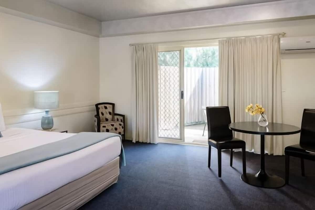 Habitación de hotel con cama, escritorio, dos sillas, una mesita con flores y puerta corrediza de vidrio al exterior.