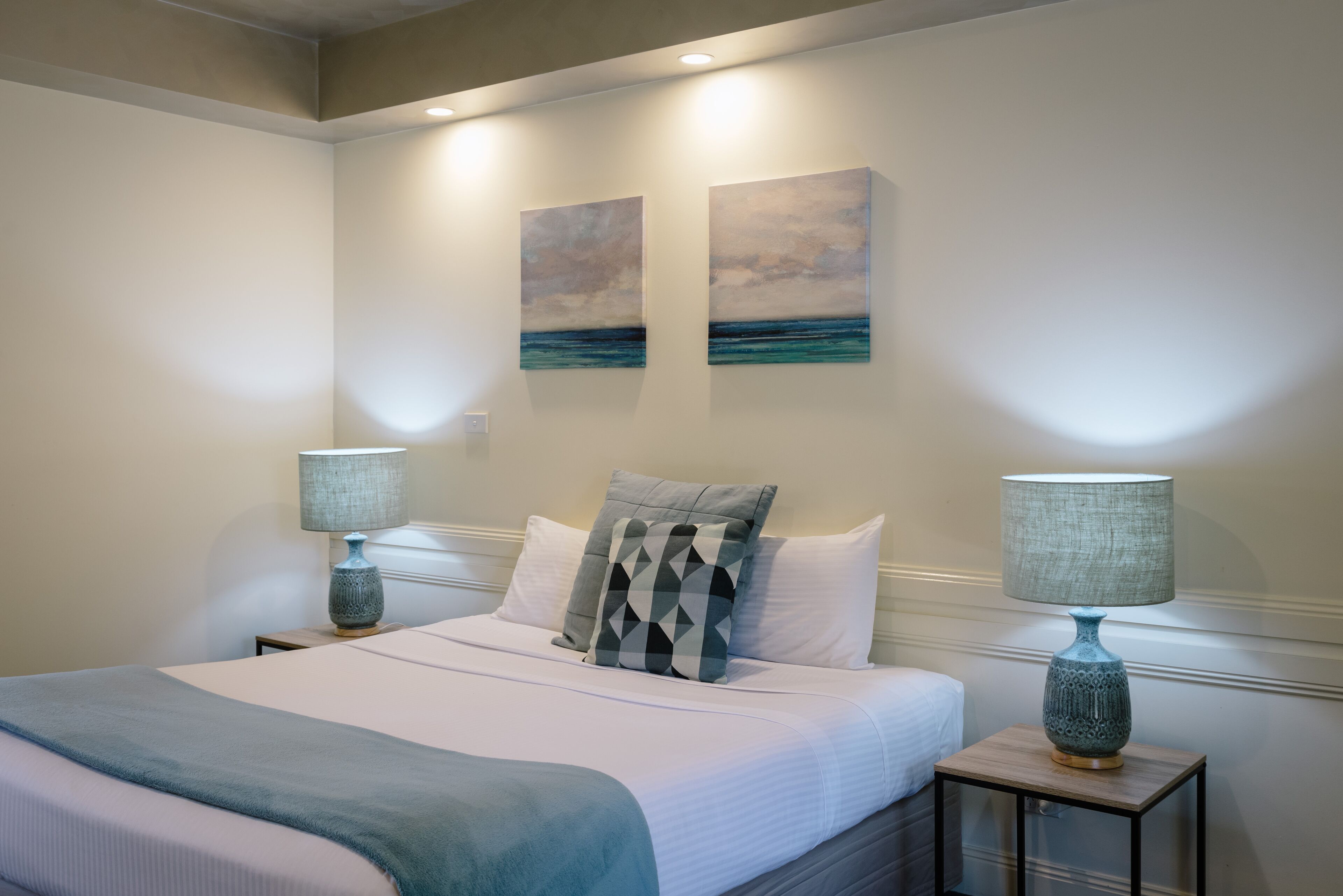 A neatly made bed with a blue blanket, two matching lamps, and two paintings on the wall.