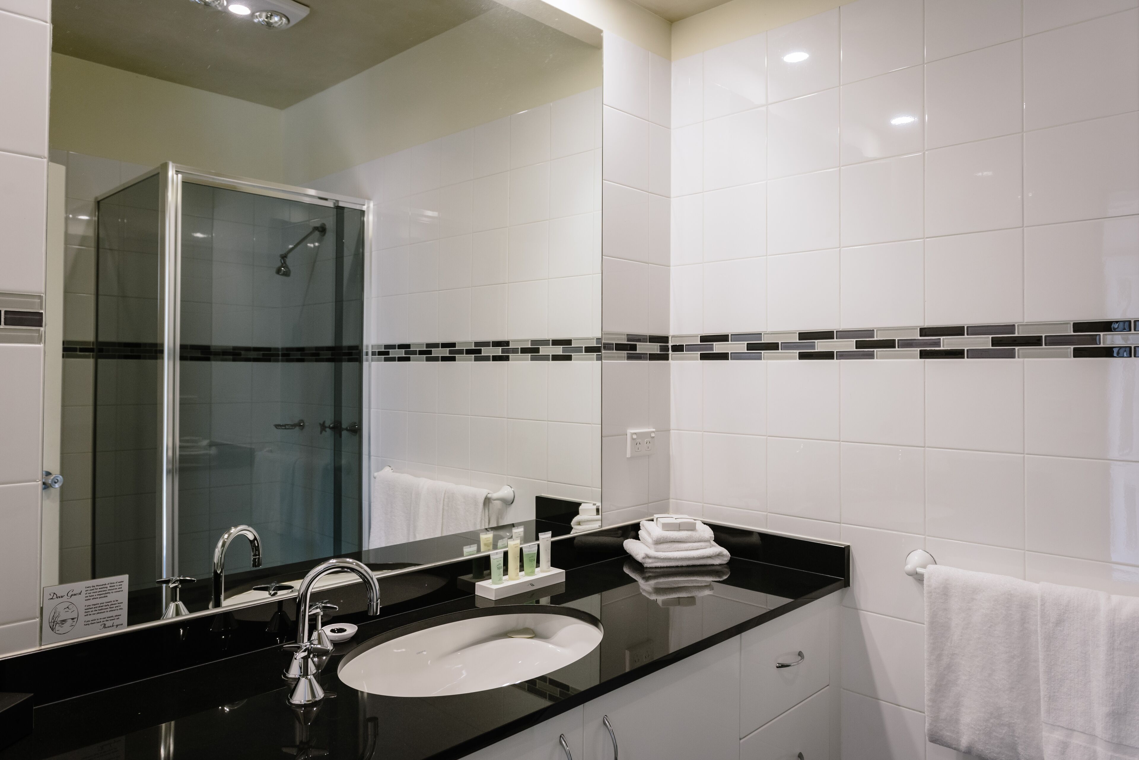 Une salle de bain moderne avec une douche à parois de verre, un lavabo sur un plan de toilette noir et des murs recouverts de carreaux blancs.