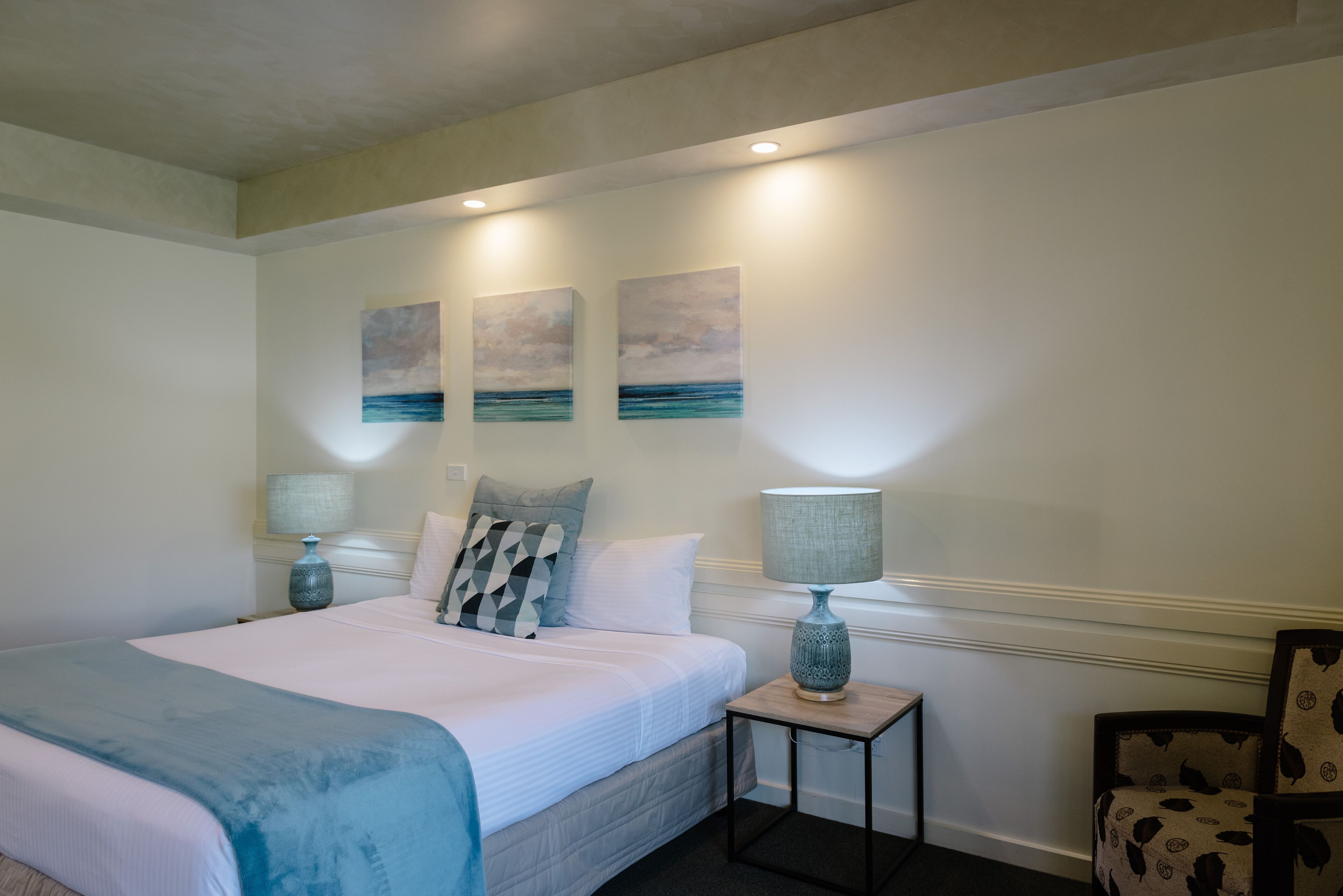 A bedroom with a large bed, two bedside tables with lamps, a chair, and three wall-mounted paintings depicting a seascape.