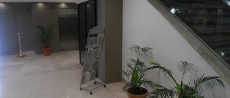 A modern lobby with a glass staircase, potted plants, and a metal information stand.