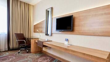 Habitación de hotel con escritorio de madera, una silla, televisor montado en la pared y un ventanal con cortinas.