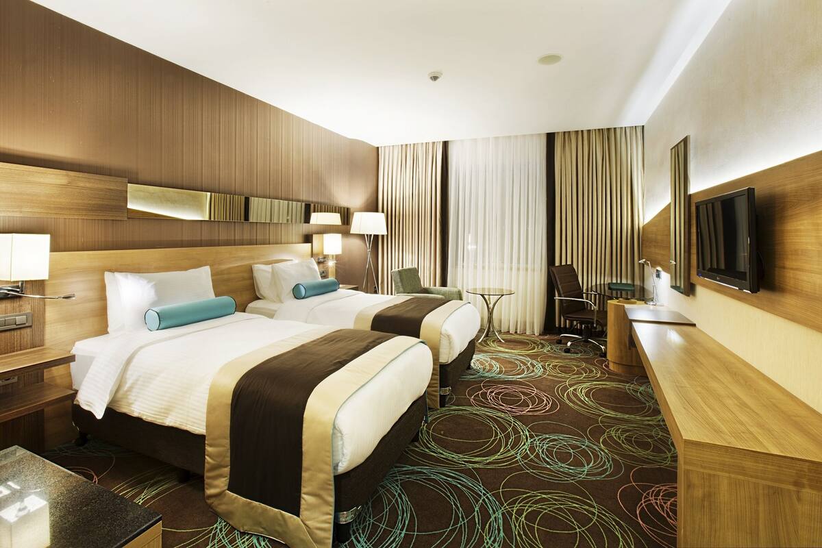 A hotel room with two beds, a desk, a chair, a TV, and a window with curtains.
