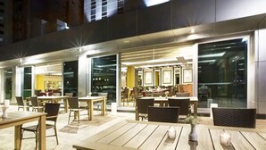 Un área de comedor al aire libre con mesas y sillas, ventanas grandes y vistas a la ciudad.