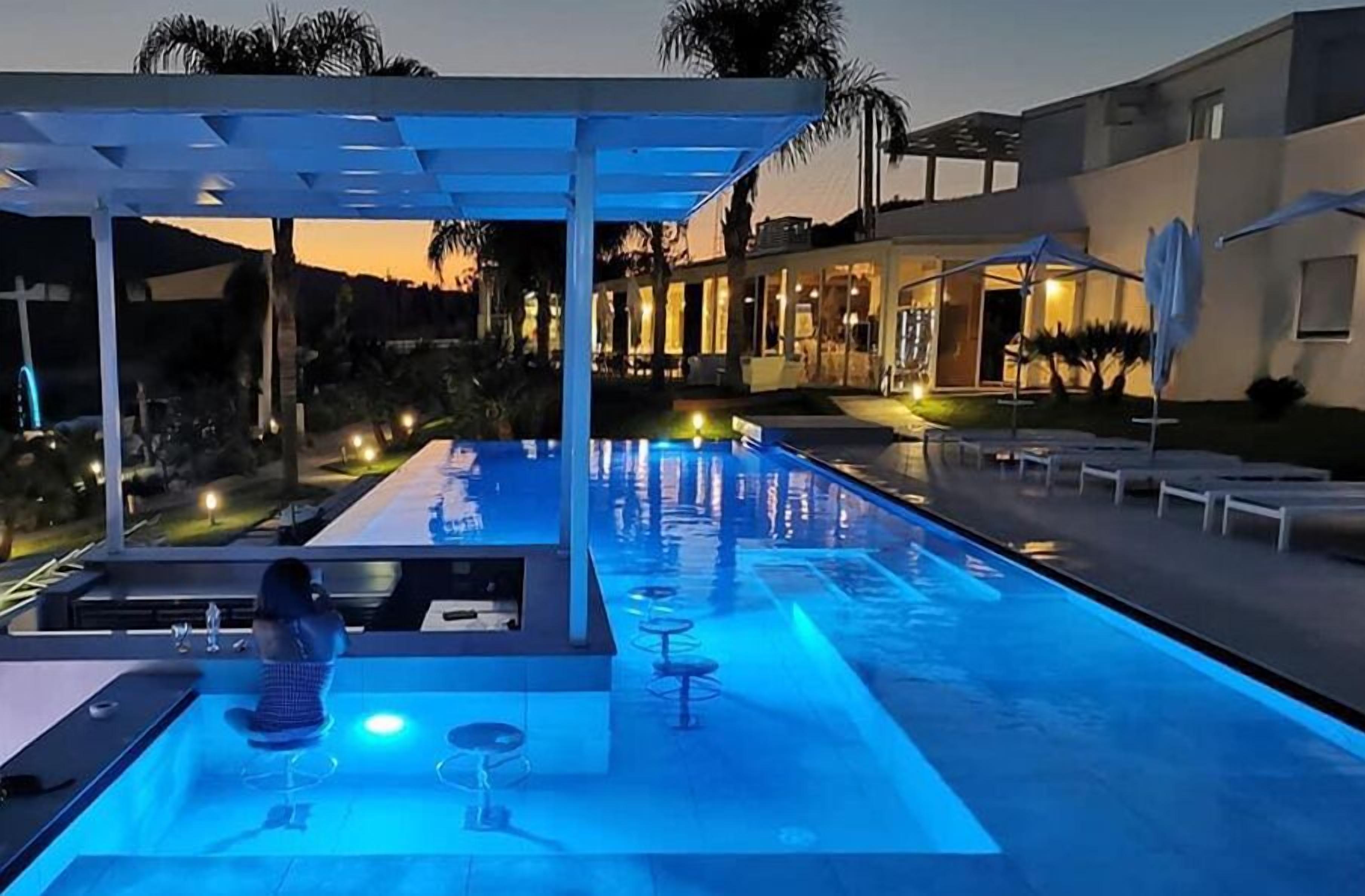 A swimming pool with a covered area and lounge chairs, surrounded by a landscaped garden and a building with illuminated windows.