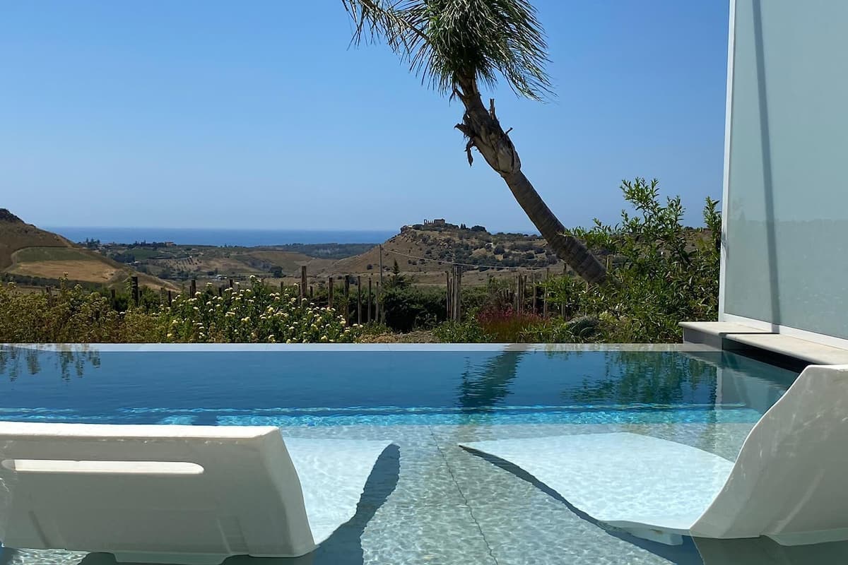 Una piscina infinita de color azul intenso con dos sillas reclinables blancas, una palmera y vistas al océano y colinas.