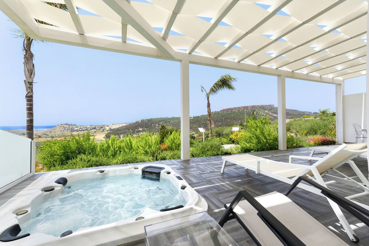 Un jacuzzi con vista al océano y colinas, rodeado de sillas de descanso y una pérgola.