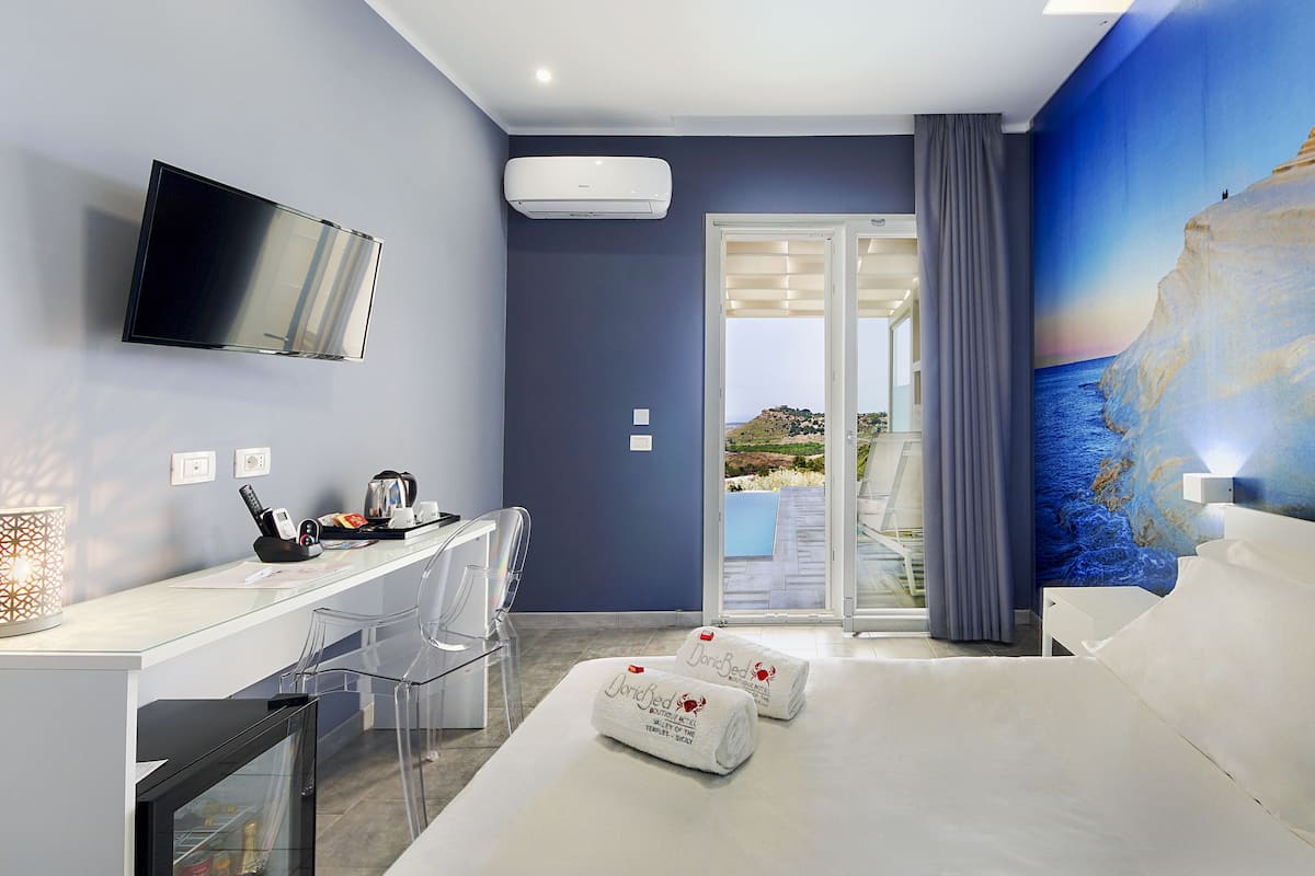 Habitación de hotel con cama, escritorio con televisor de pantalla plana, cafetera y vista al mar a través de un ventanal.