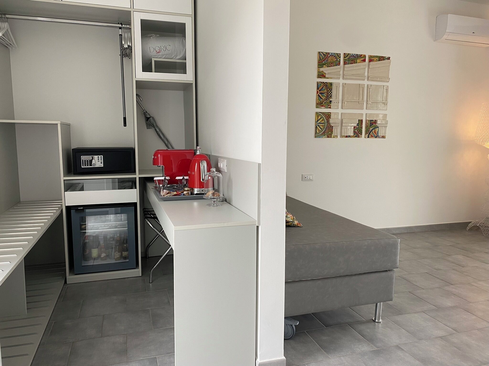 Un espacio habitable compacto con kitchenette, un sofá gris y un aire acondicionado montado en la pared.
